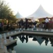 Wedding reception in progress next to a pool with a Hexagon 100 wedding tent and luminarias ready to light the deck.