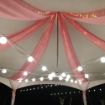 A Hexagon 60 wedding tent with pink canopy swagging highlighted by twinkle lights and elegant globe lighting.